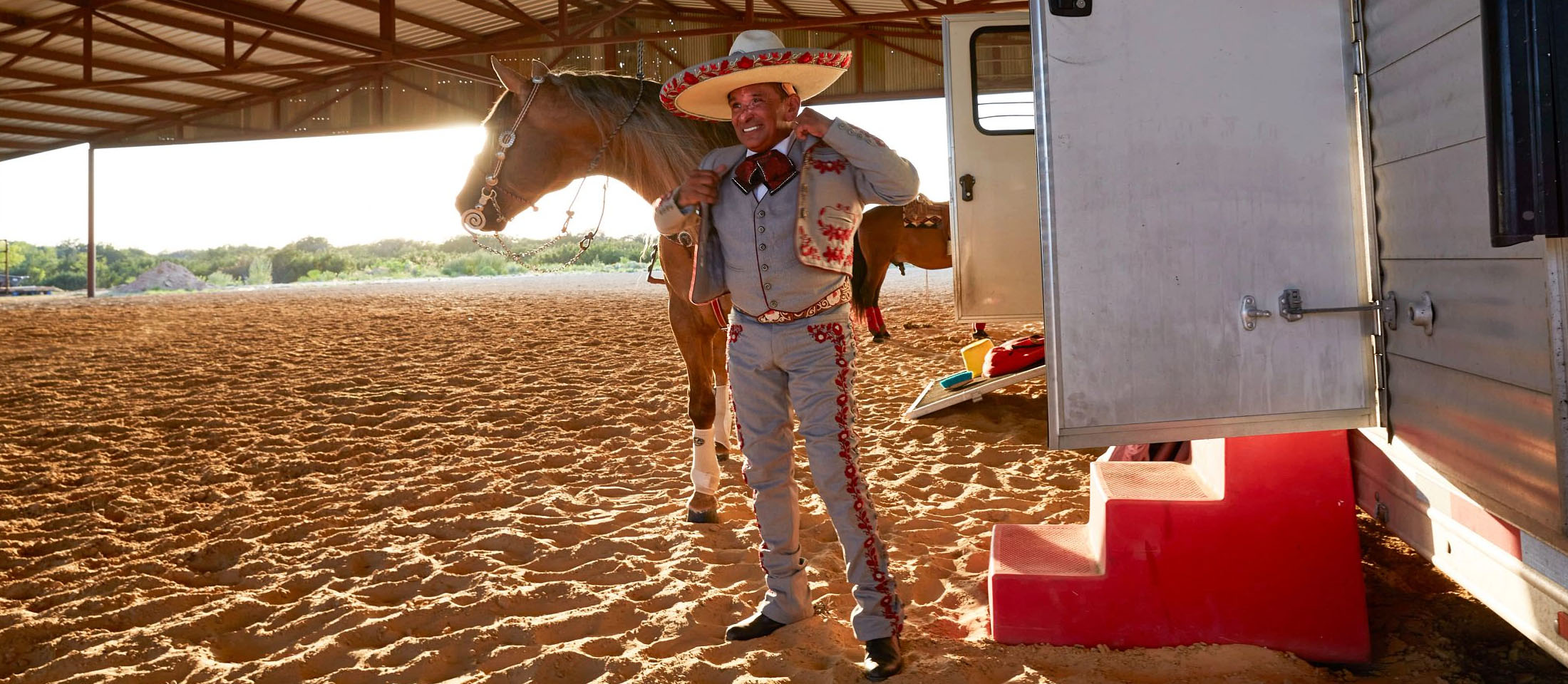 Un hombre vestido con sombrero y traje tradicional de Charro poniéndose una chaqueta frente a un caballo y de pie junto a un remolque para caballos.